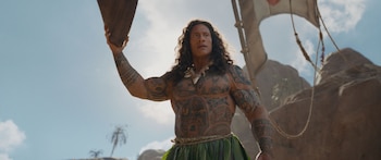 Dwayne Johnson, con cabello largo y rizado, luce tatuajes tribales en torso y brazos, un collar de dientes y una falda de hojas verdes, sosteniendo un remo bajo un cielo azul