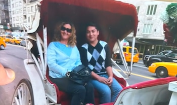 Una mujer y un joven sentados en el interior de un carruaje blanco con capota roja en una avenida con automóviles amarillos