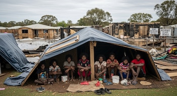 Ocho jóvenes indígenas sentados dentro de un refugio de lona azul. Detrás, casas inundadas y quemadas; se aprecian letreros de alquiler y seguro denegado.