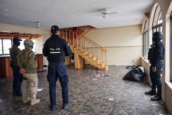 Cuatro hombres con vestimenta táctica en el interior de una propiedad con piso de baldosas, escaleras de madera, ventanas arqueadas y un cubo de basura negro.
