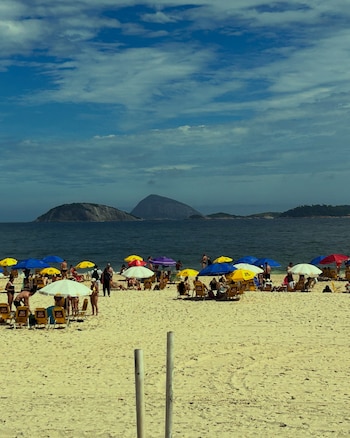 Sombrillas de colores, reposeras y personas en una playa de arena frente al mar, con islas montañosas al fondo bajo un cielo nublado