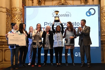 Un grupo de personas sobre un escenario; una mujer sostiene un trofeo en alto y otra muestra un diploma junto a un hombre en traje que aplaude