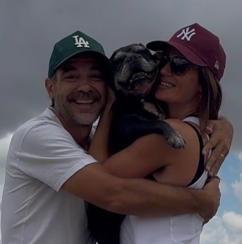 Paula Chaves y Pedro Alfonso sonríen mientras abrazan a un perro de raza pug de pelaje oscuro. Llevan gorras y ropa clara bajo un cielo nublado