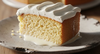 Primer plano de una porción de pastel tres leches sobre un plato rústico, mostrando su textura aireada, glaseado blanco con gotas de humedad y leche derramada.