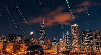 Vista nocturna de los rascacielos de Chicago bajo un cielo oscuro, con múltiples trazas de meteoros rojos y verdes cruzando el firmamento.