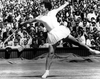 Imagen en blanco y negro de Billie Jean King en una cancha de tenis, vestida de blanco, realizando un golpe con la raqueta, con público al fondo