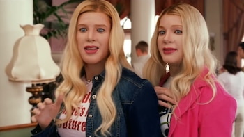 Dos personas con pelucas rubias, maquillaje exagerado, y ojos azules grandes miran sorprendidas. Una lleva chaqueta vaquera, la otra un blazer rosa
