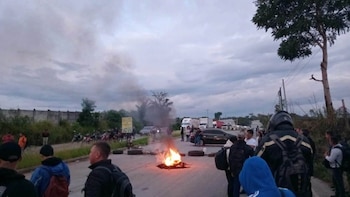 Personas reunidas alrededor de un fuego en el asfalto de una carretera, con camiones y automóviles detenidos al fondo