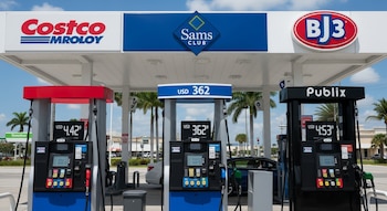 Tres surtidores de gasolina bajo un toldo con logos de Costco, Sam's Club y BJ's. Muestran precios USD 4,42, USD 3,62 y USD 4,53. Fondo urbano.