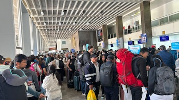 Numerosas personas en una fila larga dentro de un aeropuerto con maletas y mochilas. Se observan mostradores de facturación y carteles de aerolíneas