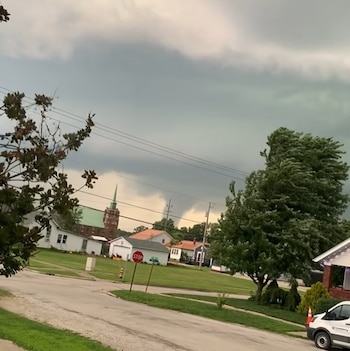 Cielo oscuro con nubes de tormenta sobre un barrio. Se observa una columna de tornado, casas, árboles, un campanario, cables y una camioneta