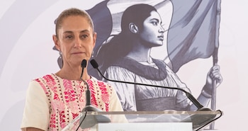 Claudia Sheinbaum detrás de un atril con micrófonos frente a un mural con la ilustración de una mujer que sostiene una bandera