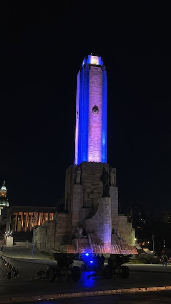 Fotografía nocturna del Monumento Nacional a la Bandera en Rosario, Argentina, con su torre principal iluminada en azul y blanco, y cañones en la base