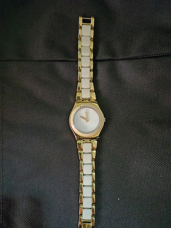 Primer plano de un reloj analógico con caja dorada y esfera clara, unido a una correa de eslabones alternos dorados y blancos sobre un fondo oscuro