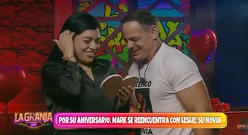 Mark Vito, en camiseta blanca, y Leslie, en camisa negra, sonríen mientras él sostiene un objeto con forma de corazón. Globos rojos adornan el fondo