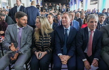 Cuatro personas sentadas en una fila durante un evento concurrido, vestidas con trajes formales y chaquetas, rodeadas por un auditorio lleno