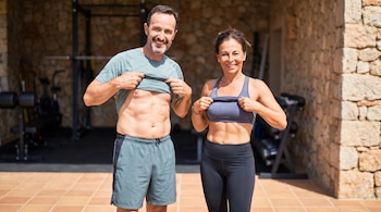 Un hombre y una mujer de mediana edad, ambos sonrientes, levantan sus camisetas para mostrar abdominales tonificados. Están de pie frente a un gimnasio al aire libre.