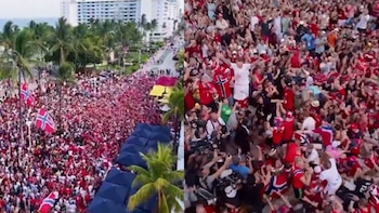 Vista aérea de una gran multitud de personas, algunas con banderas noruegas, en una calle urbana con palmeras y edificios al fondo