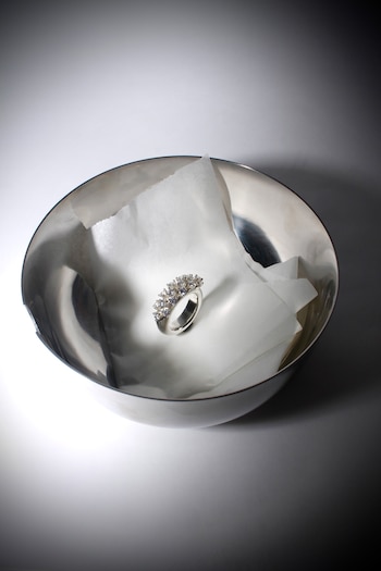 Un anillo de plata brillante con gemas transparentes descansa sobre papel arrugado dentro de un cuenco metálico pulido con fondo claro