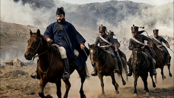 Cinco hombres con uniformes militares de época y sombreros montan caballos oscuros sobre terreno rocoso y polvoriento bajo un cielo nublado, con montañas al fondo