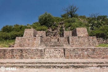 Estructuras arqueológicas de piedra con escalones y plataformas en una colina, vegetación verde y un cielo azul claro. Logotipo de INAH