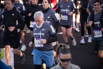 Un hombre de barba blanca y camiseta gris corre en una calle asfaltada junto a otros atletas con dorsales numerados