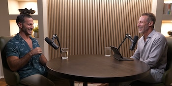 Dos hombres sentados en sillas frente a una mesa redonda de madera con dos micrófonos, dos vasos de agua y una tableta. Fondo con paneles de madera y estantes