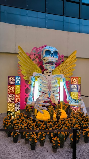 Figura de un esqueleto con alas doradas, decorado con patrones azules, rodeado por macetas de flores amarillas y columnas iluminadas