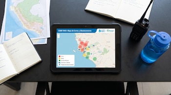 Una tableta con un mapa de zonas de corte de agua sobre un escritorio junto a libretas abiertas, un mapa impreso, una radio y una botella.