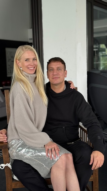 Una mujer rubia y un hombre de cabello castaño posan sonrientes. La mujer viste un jersey beige y falda plateada, sentada sobre el regazo del hombre, quien usa un buzo negro