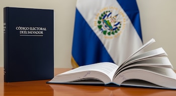 Dos libros del Código Electoral de El Salvador sobre una mesa de madera, uno abierto y uno cerrado, con la bandera de El Salvador difuminada al fondo.