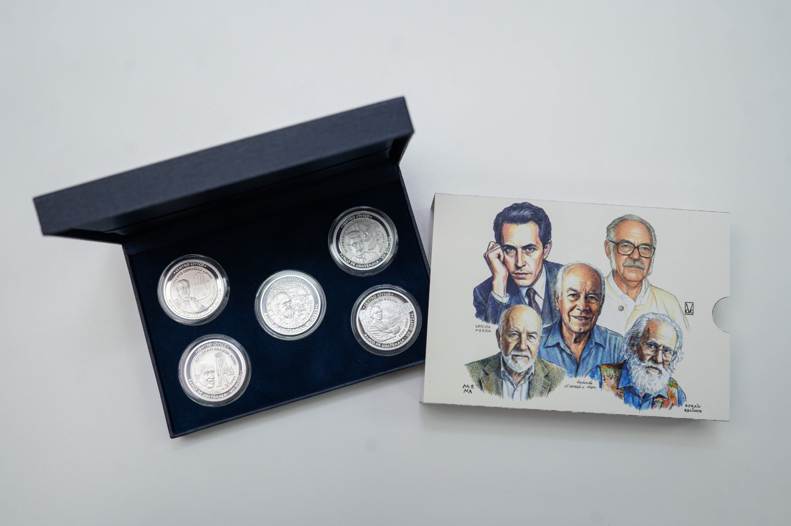 Una colección de cinco monedas conmemorativas del Banco de Guatemala, junto con un folleto ilustrado con los rostros de importantes figuras artísticas y culturales del país, se exhiben sobre una superficie blanca. (Banco de Guatemala)
