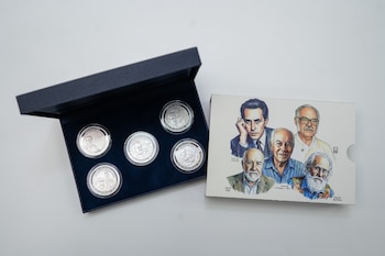Cinco monedas conmemorativas de plata en su caja abierta y un folleto ilustrado con cinco retratos de hombres, sobre un fondo liso y claro