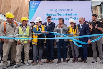 Un grupo de trabajadores con cascos de seguridad y personas con traje cortan una cinta azul frente a una pantalla informativa