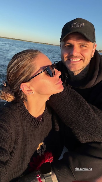 Mujer y hombre en primer plano, con agua y cielo azul de fondo. La mujer usa gafas de sol y suéter negro; el hombre, gorra y sudadera negras