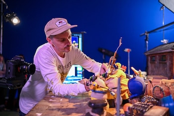 Un hombre con gorra ajusta varias figuras pequeñas sobre una mesa de madera junto a una cámara fotográfica profesional