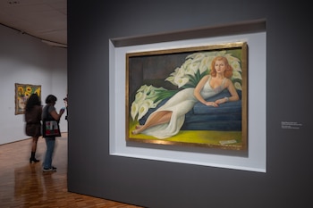 Pintura de una mujer rubia recostada entre alcatraces blancos en un museo, con dos personas observando otras obras al fondo