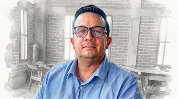 Hombre con camisa azul y gafas, frente a una pared de ladrillo y ventanas. El fondo está difuminado con efecto acuarela en tonos grises.