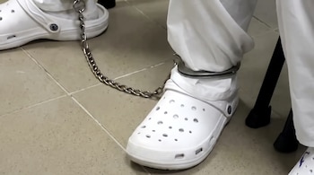 Dos pies con zapatos blancos tipo zueco, tobilleras metálicas con cadena que los une, pantalones blancos y suelo de baldosas.