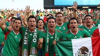 Aficionados del fútbol mexicano en primer plano, con camisetas verdes, bufandas y cara pintada, gritando y levantando los brazos. Uno sostiene una bandera de México.