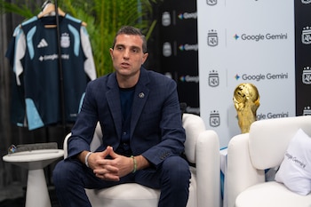 Leandro Petersen, con traje azul, sentado hablando en un evento. Detrás, camiseta de Argentina, trofeo dorado y logos de Google Gemini y AFA