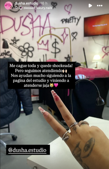 Primer plano de una mano con uñas acrílicas decoradas con patrones y la palabra 'Baby', anillos y un tatuaje de ojo en el dorso. Fondo de pared con grafitis