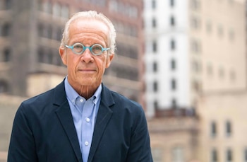 Retrato de un hombre de cabello canoso con chaqueta oscura, camisa azul y gafas redondas celestes, frente a edificios desenfocados