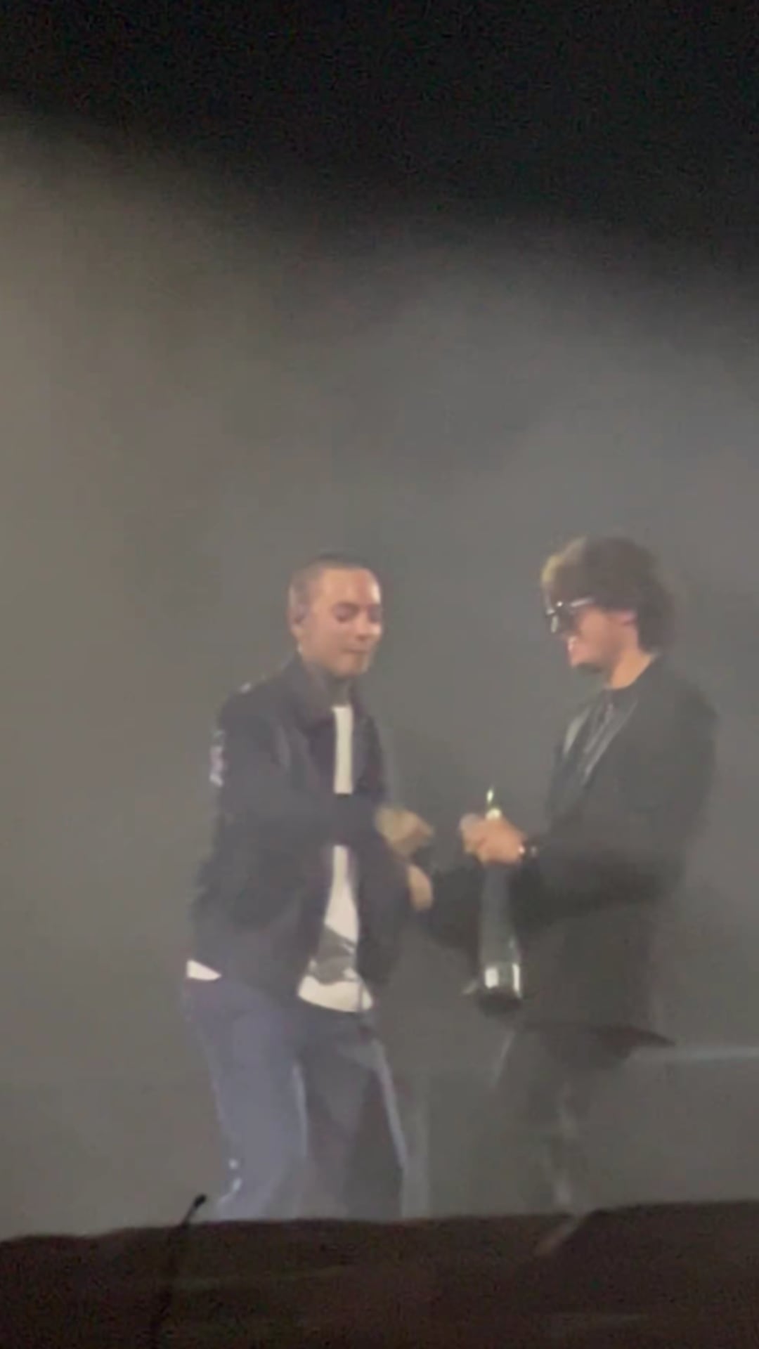 Los cantantes Natanael Cano y Gabito Ballesteros comparten el escenario y sostienen una botella durante una presentación musical en vivo. (X)