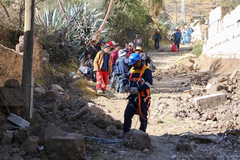 Un trabajador con casco azul y arnés en un camino lleno de escombros. Un grupo de personas y paredes derrumbadas se ven al fondo