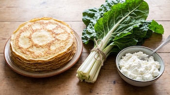 Panqueques apilados en un plato, un atado de acelga fresca y un recipiente con ricota y una cuchara sobre una mesa de madera.