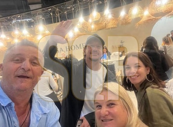 Selfie de cuatro personas sonriendo. Nico Vázquez de gorra y chaqueta oscura, saludando. Dai Fernández a la derecha, sonríe con aros. Fondo con luces de mimbre