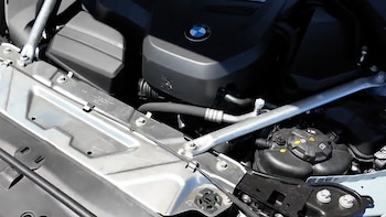 Vista cercana del compartimento del motor de un automóvil, con el logo de BMW visible, tuberías, cables y una cubierta del motor con signos de deterioro