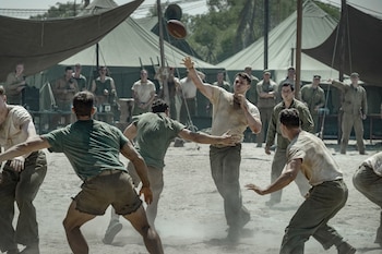 Varios hombres vestidos con prendas militares juegan al fútbol americano sobre tierra, con carpas militares y espectadores al fondo