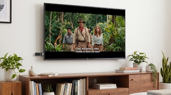 Televisor negro en pared blanca reproduce película con subtítulos, conectado a una memoria USB negra. Abajo hay un mueble con libros y plantas.